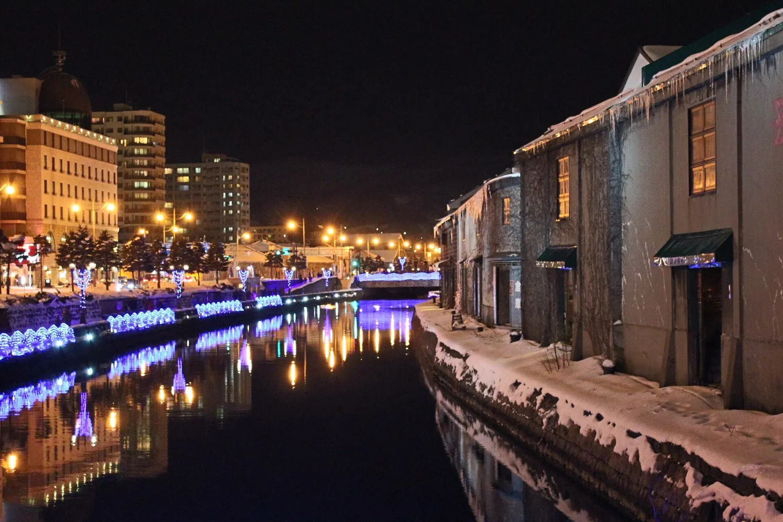 The Otaru canal at night in winter, with lit stone warehouses and snow along the water's edge
